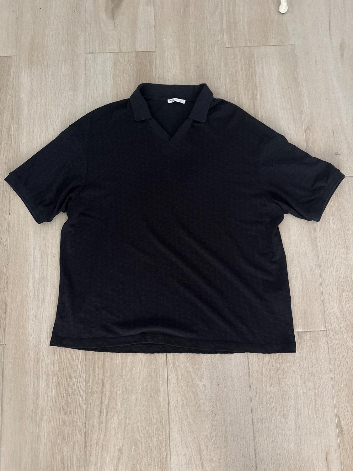 Black short-sleeved V-neck T-shirt, laid on a light wooden floor. Visible label on the collar.