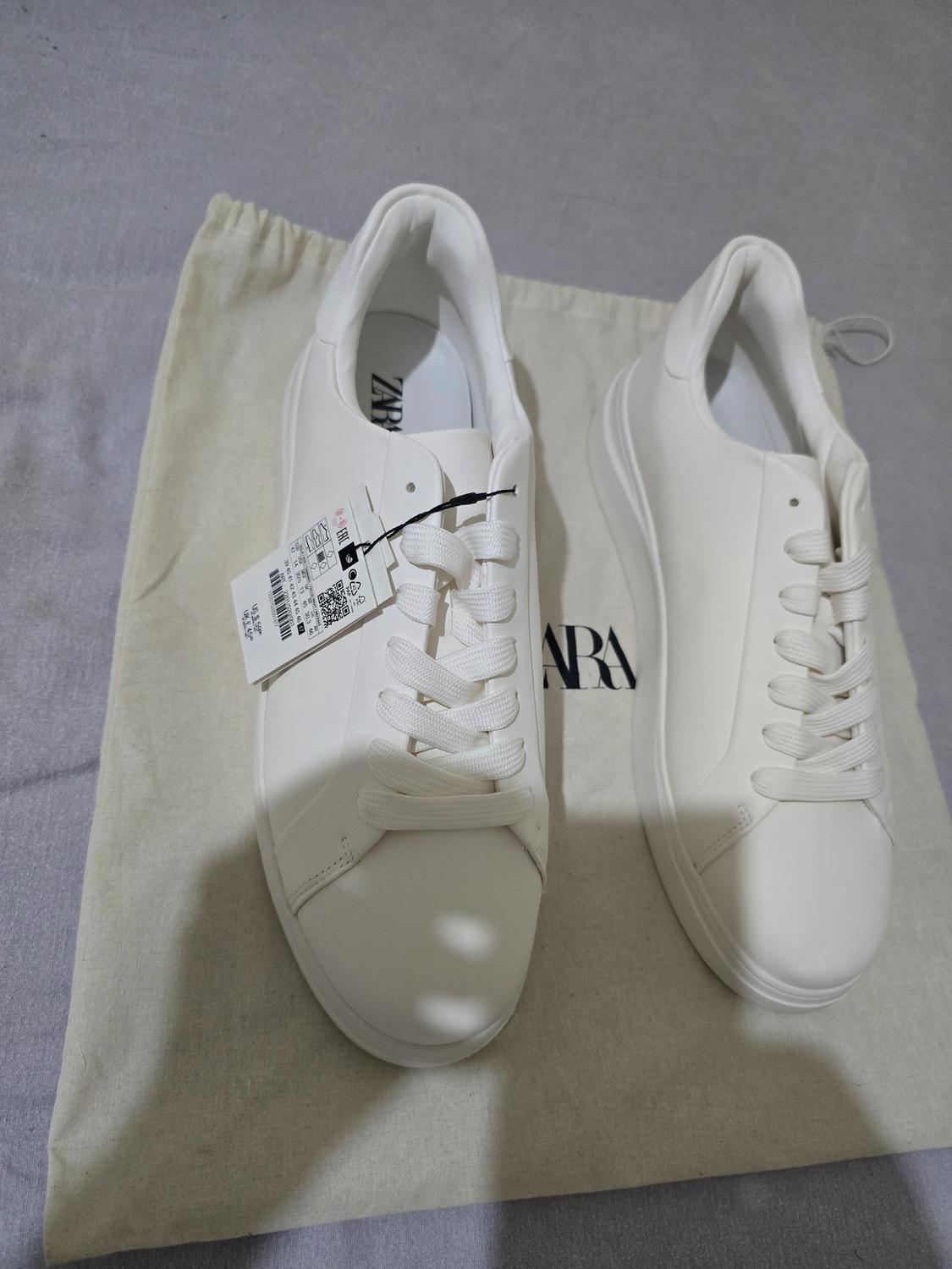 Pair of white trainers with laces and a thick sole. One has a label. They are resting on a light-coloured fabric bag with the text ZARA.