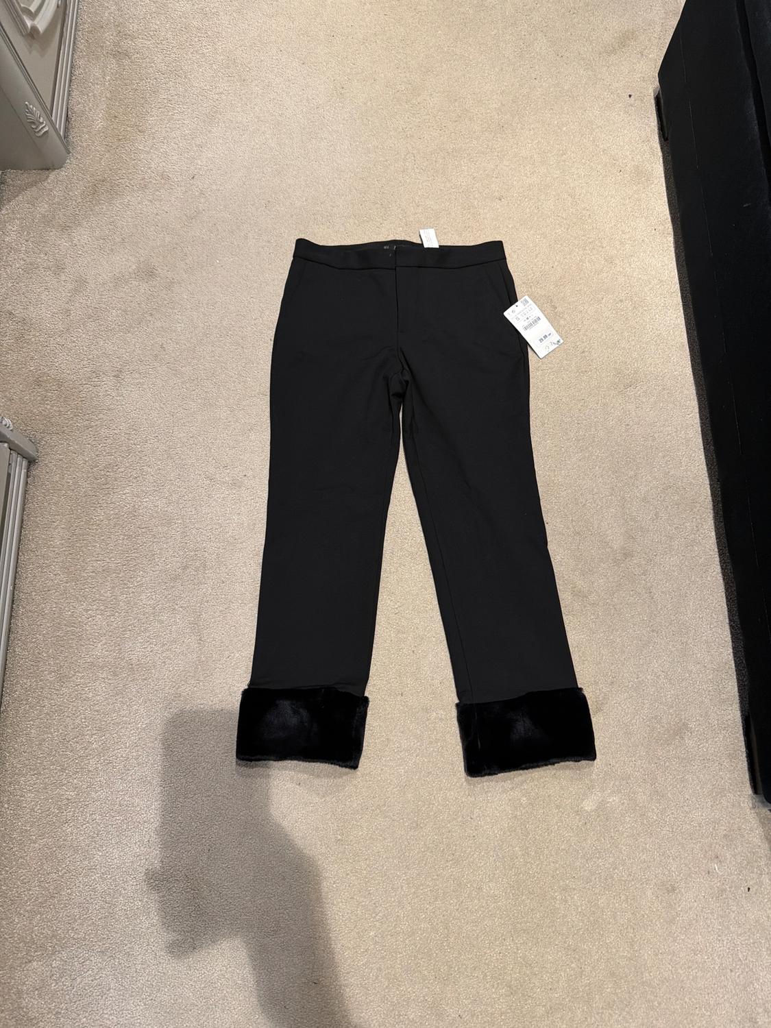 Black trousers with a faux fur hem on a shop floor, with visible tags. Beige carpet background.