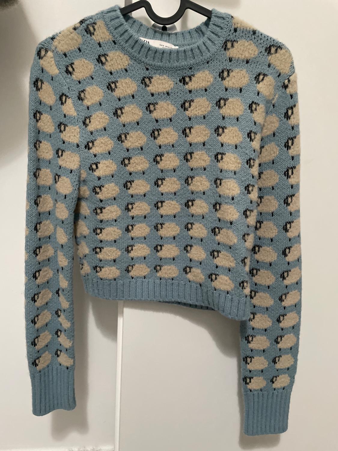 Blue jumper with beige and black sheep print, hanging on a black hanger against a white background.