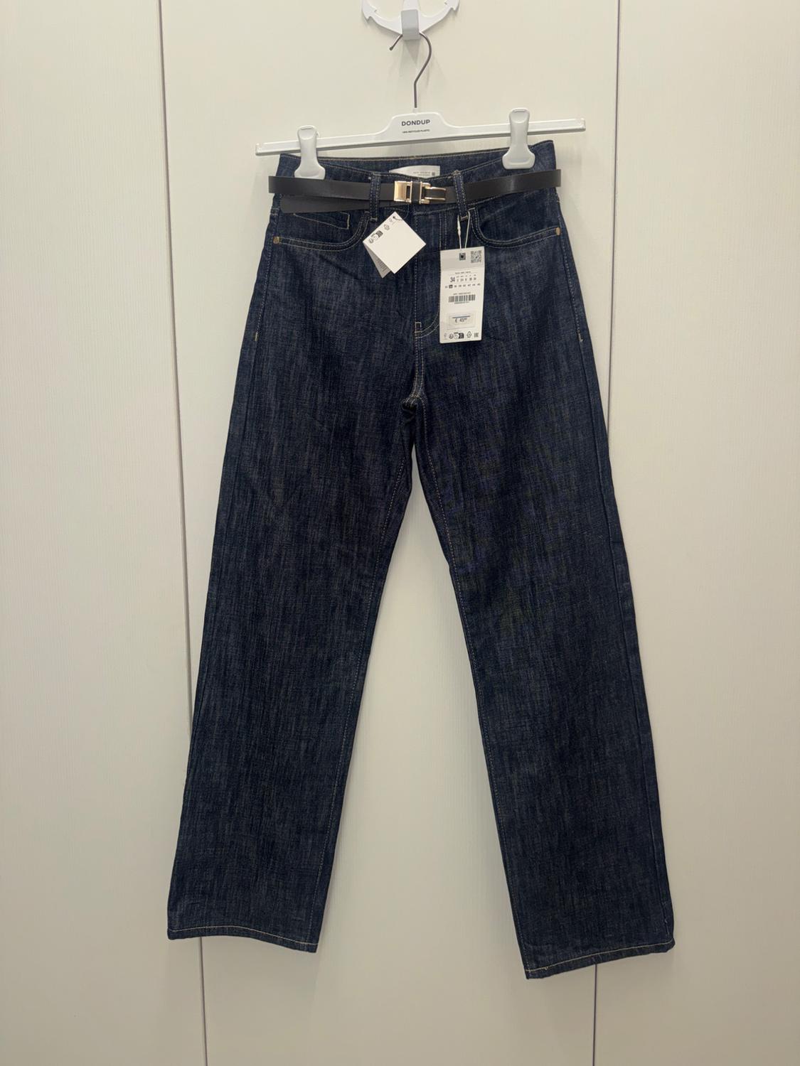 Dark denim jeans hanging on a white hanger with a black belt and visible tags. Plain white background.