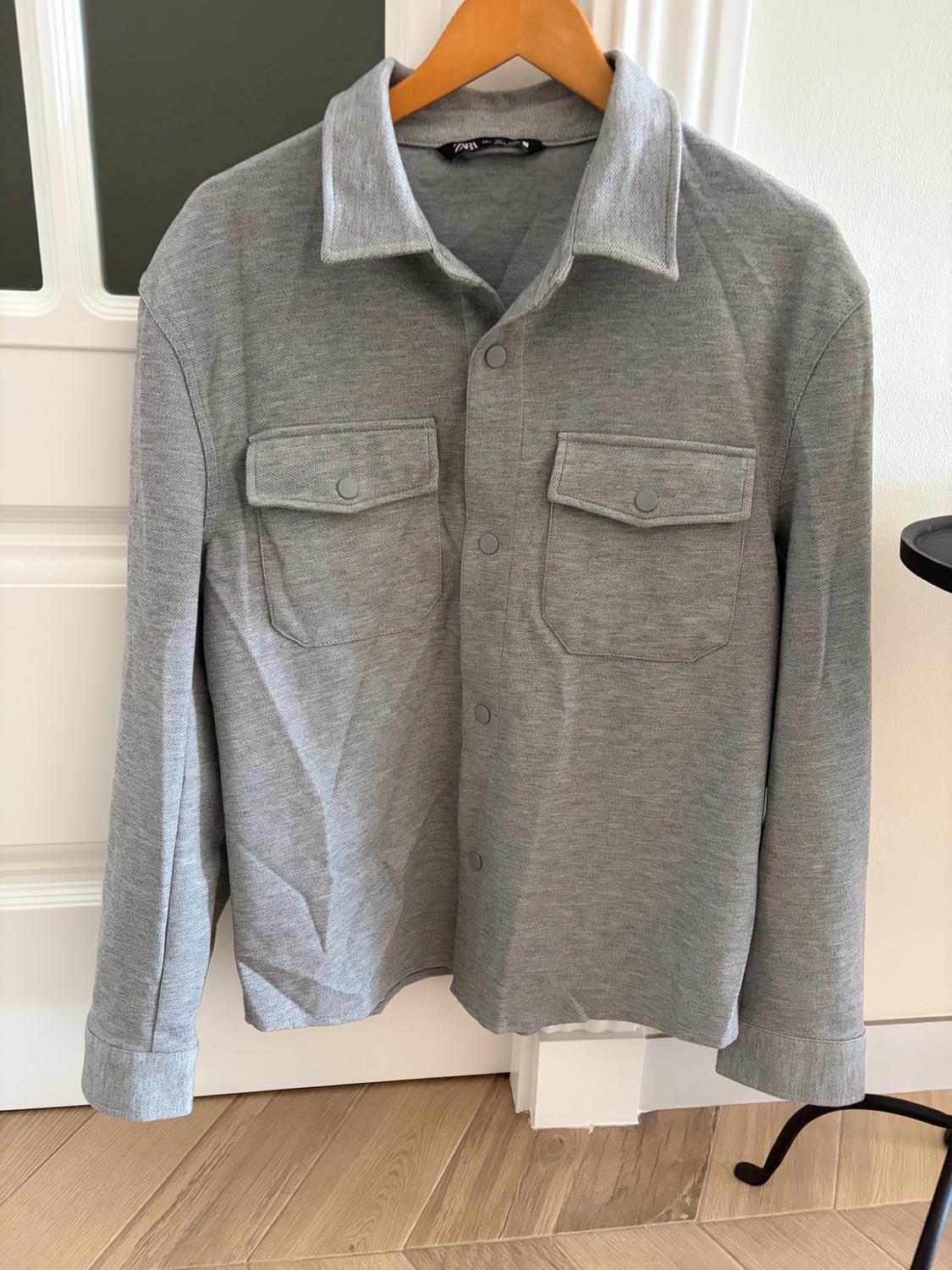 Grey long-sleeved overshirt with front pockets and ZARA label, hanging on a wooden hanger in front of a white door.