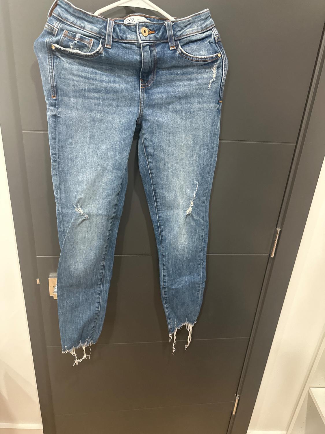 Worn blue jeans hanging on a hanger over a gray door. They have ripped details and a frayed hem.