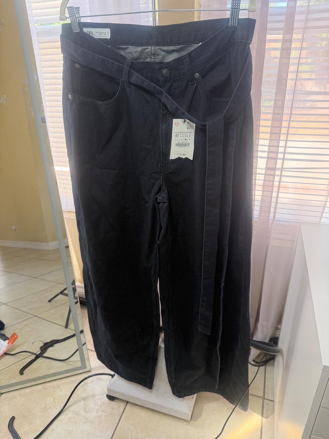 Black pants hanging on a hanger with a matching belt. Background of curtains and mirror in a brightly lit room.