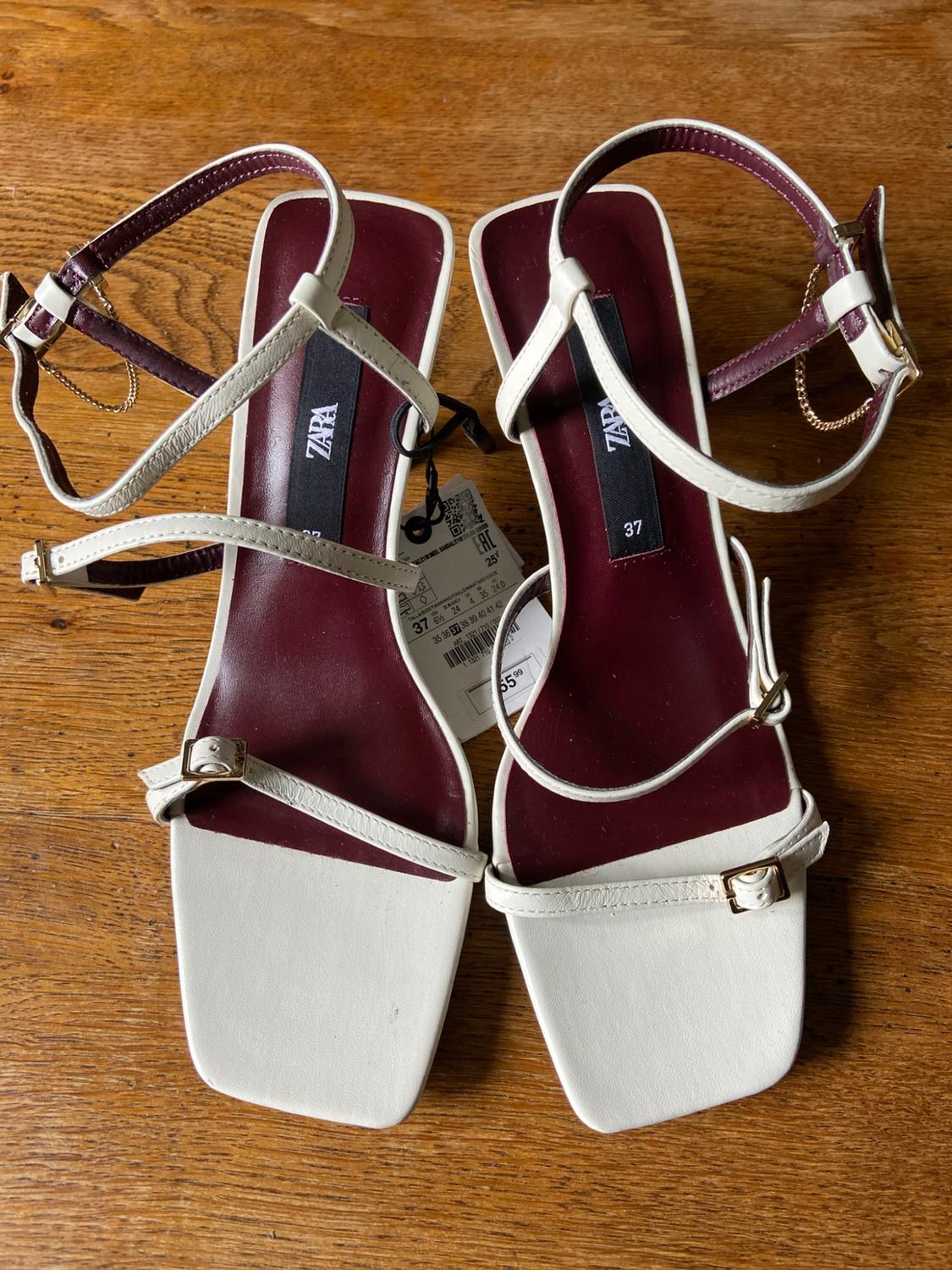 White strappy sandals with a maroon insole and gold details on a wooden surface. Text: ZARA, 37.