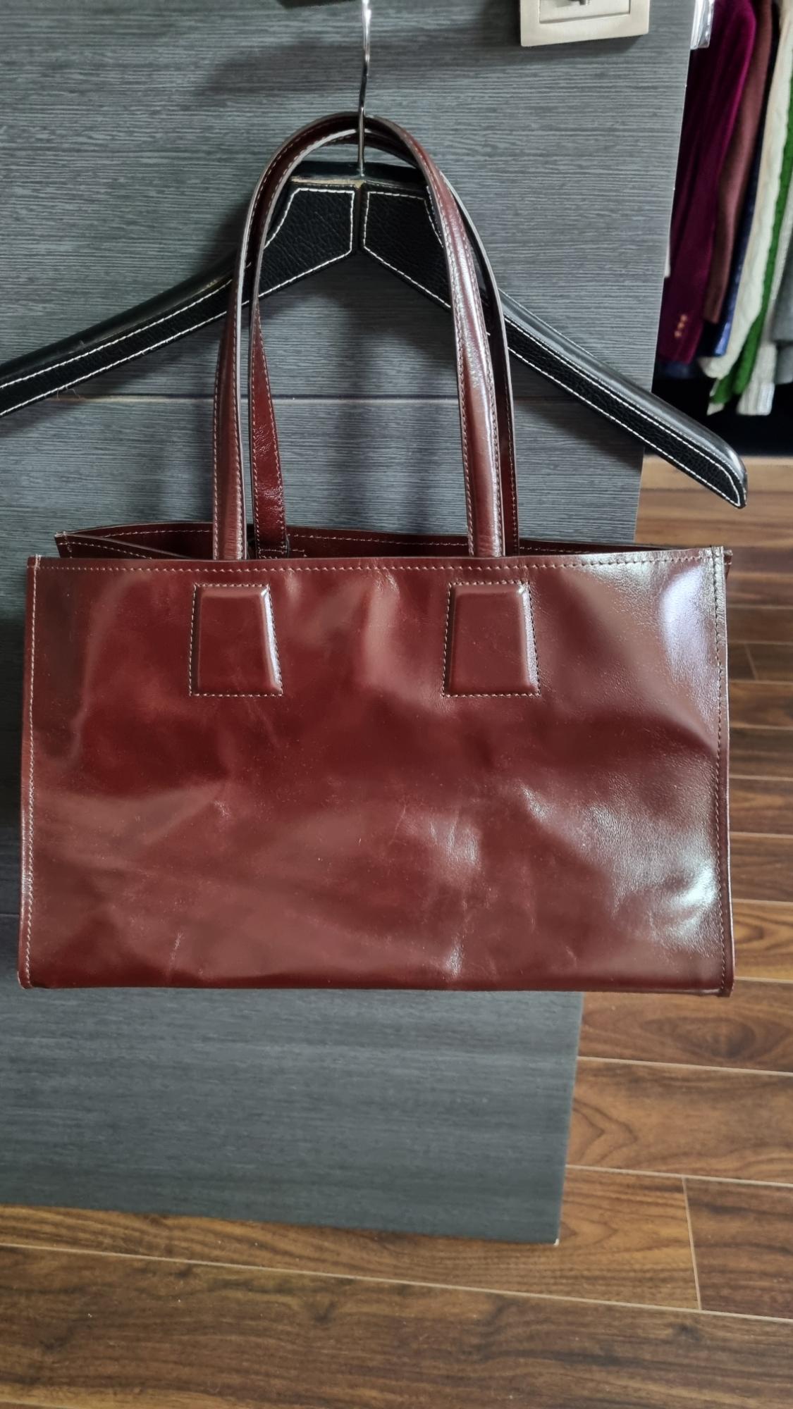 Shiny brown handbag hanging on a black coat hanger against a dark wooden background. Wooden floor visible at the bottom.