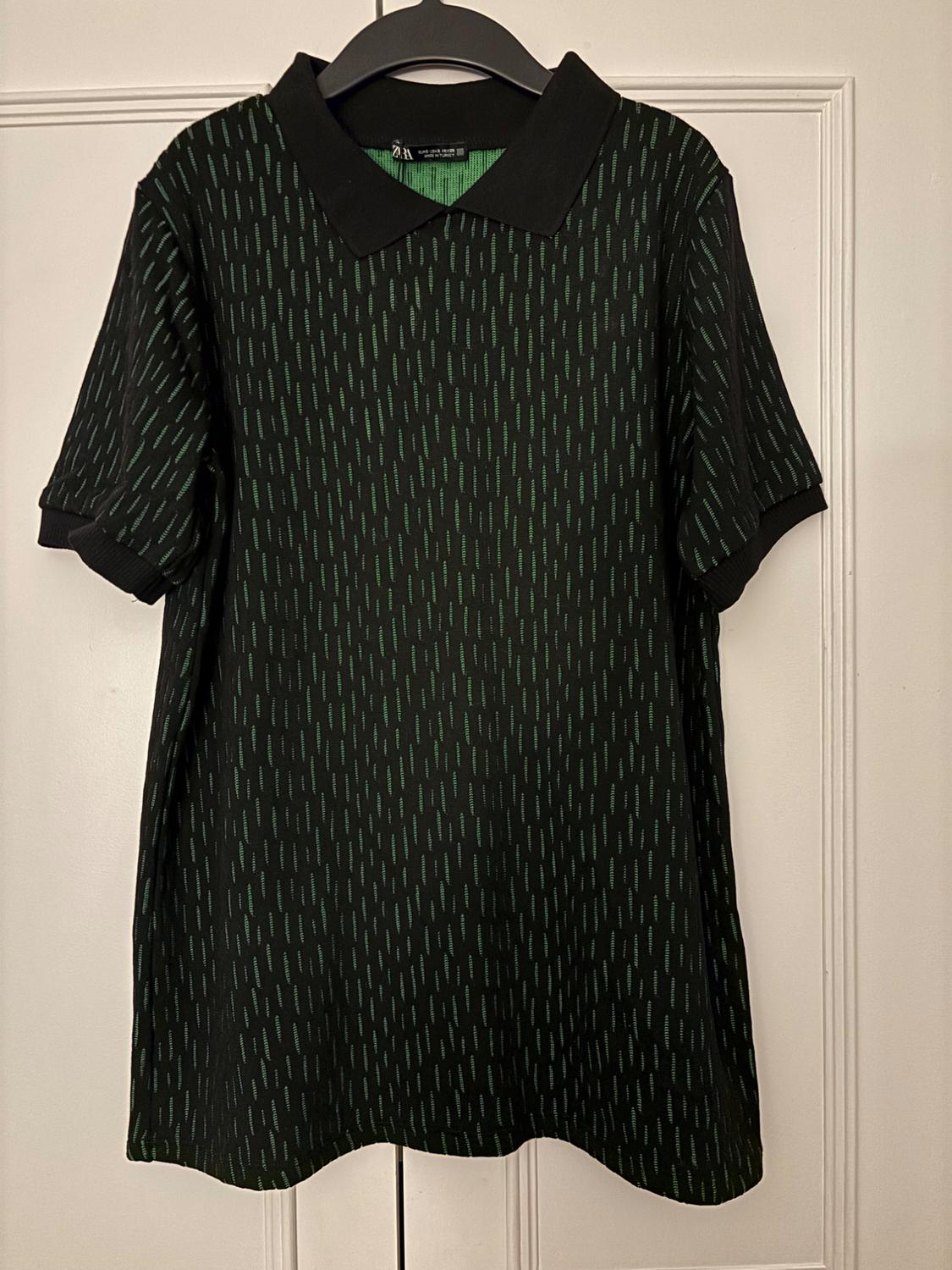Black shirt with a green line pattern, classic collar, and short sleeves, hanging on a black hanger in front of a white door.