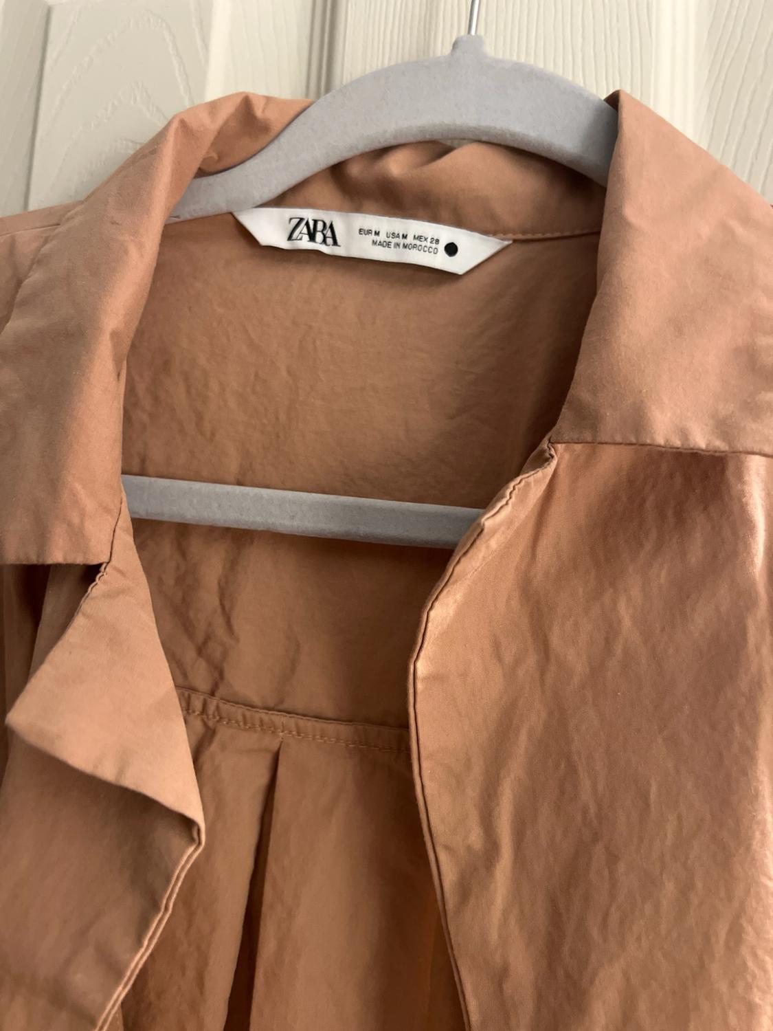 Detail of a camel-colored garment on a hanger. Label with text: ZARA EUR M USA M MEX 28 MADE IN MOROCCO.
