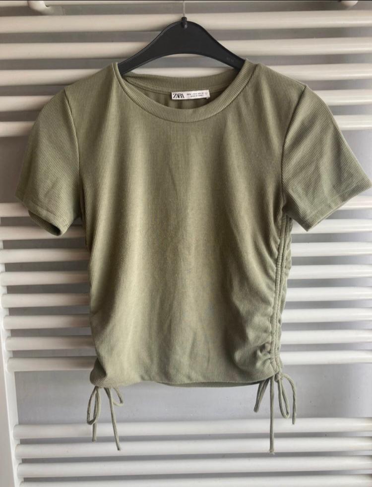 Olive green short-sleeved T-shirt with side ruching and lacing, hung on a black hanger against a white slatted background.