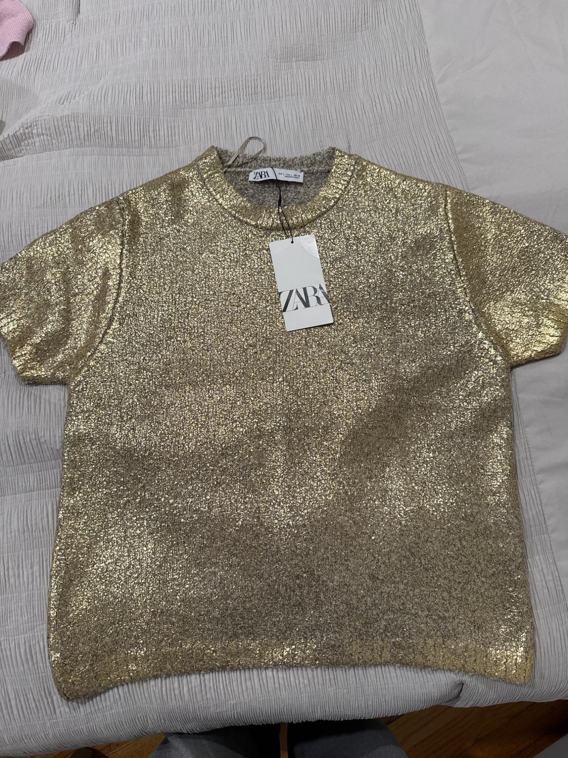Shiny gold t-shirt on a bed, with a Zara tag hanging from the collar. White and gray fabric background.