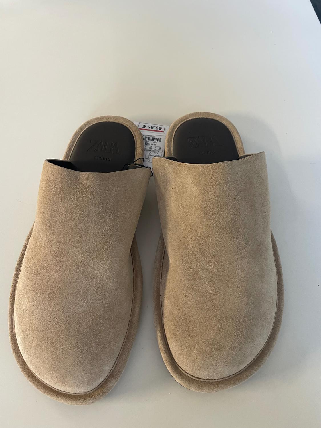 Pair of beige suede clogs on a white background. Dark insole with ZARA STUDIO text and a tag for €69.95.