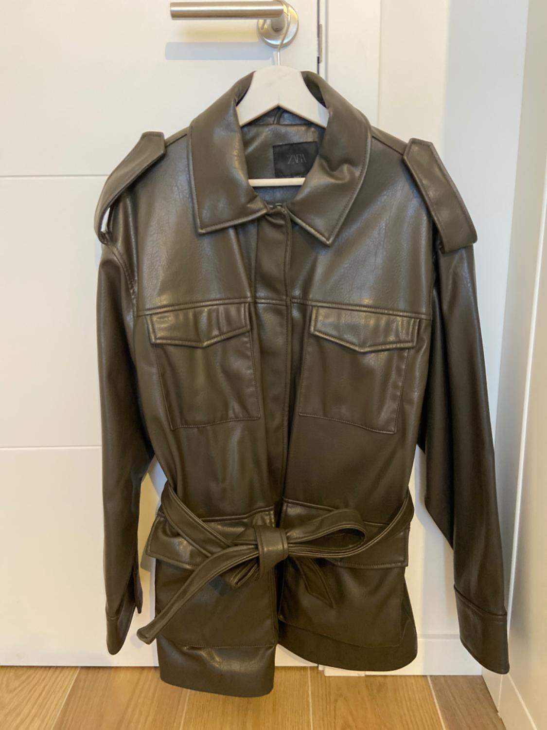 Brown faux leather jacket with belt, hanging on a white hanger in front of a white door with a metallic handle.