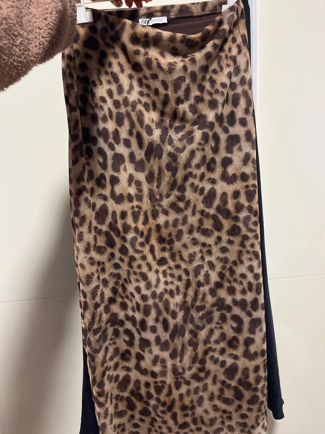 A leopard print skirt in brown and black tones, held by a hand wearing a beige jumper. The background is white.