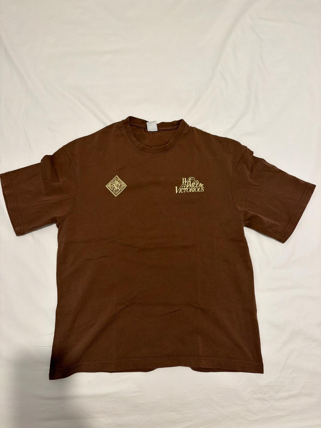 Brown short-sleeved T-shirt on white fabric. Features a diamond graphic and the text "We Will Be VICTORIOUS" on the chest.