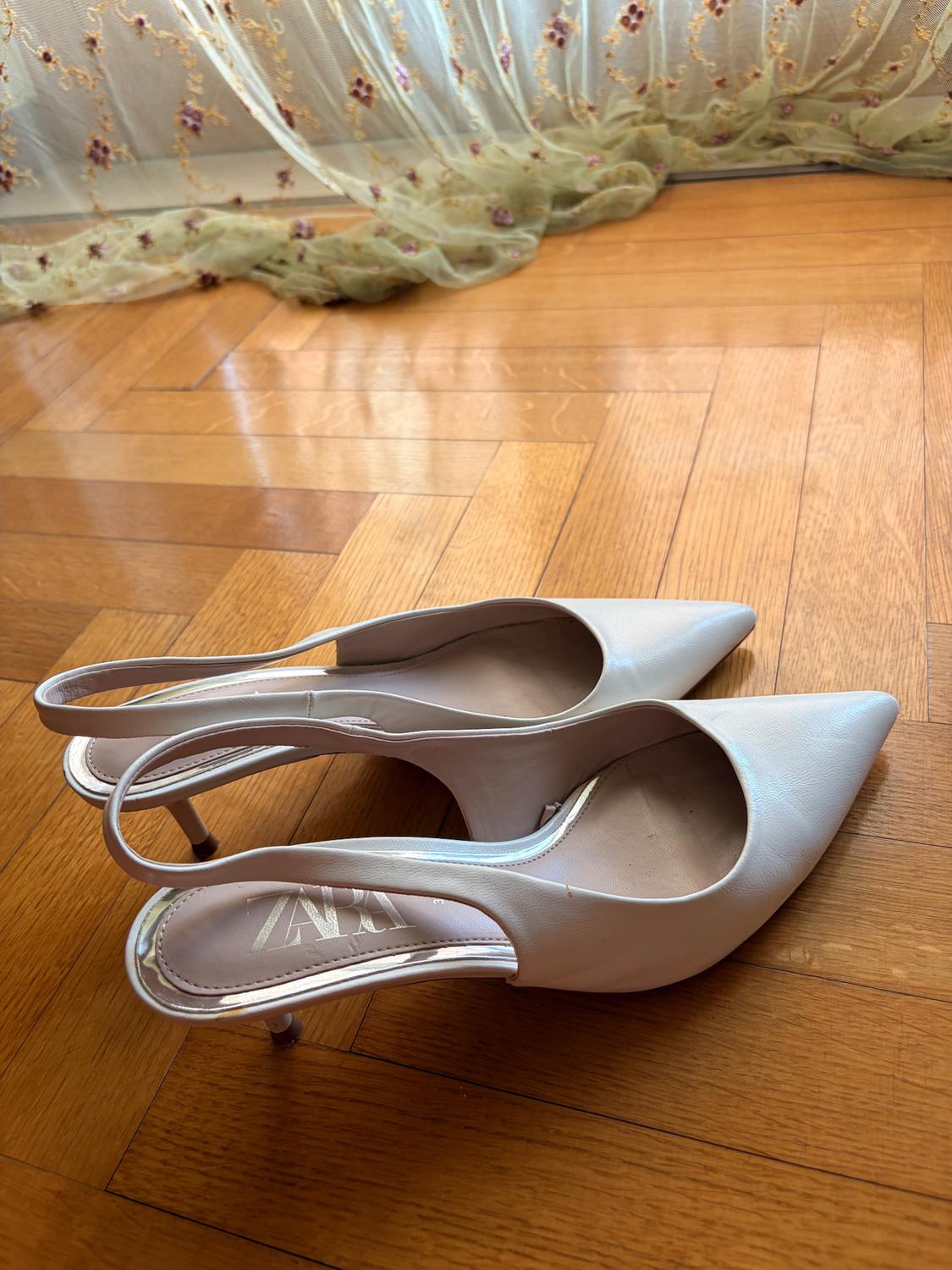Λευκά slingback παπούτσια με χαμηλό τακούνι και μυτερή σόλα σε ξύλινο δάπεδο. Στο βάθος, μια κεντημένη κουρτίνα. Κείμενο: ZARA.