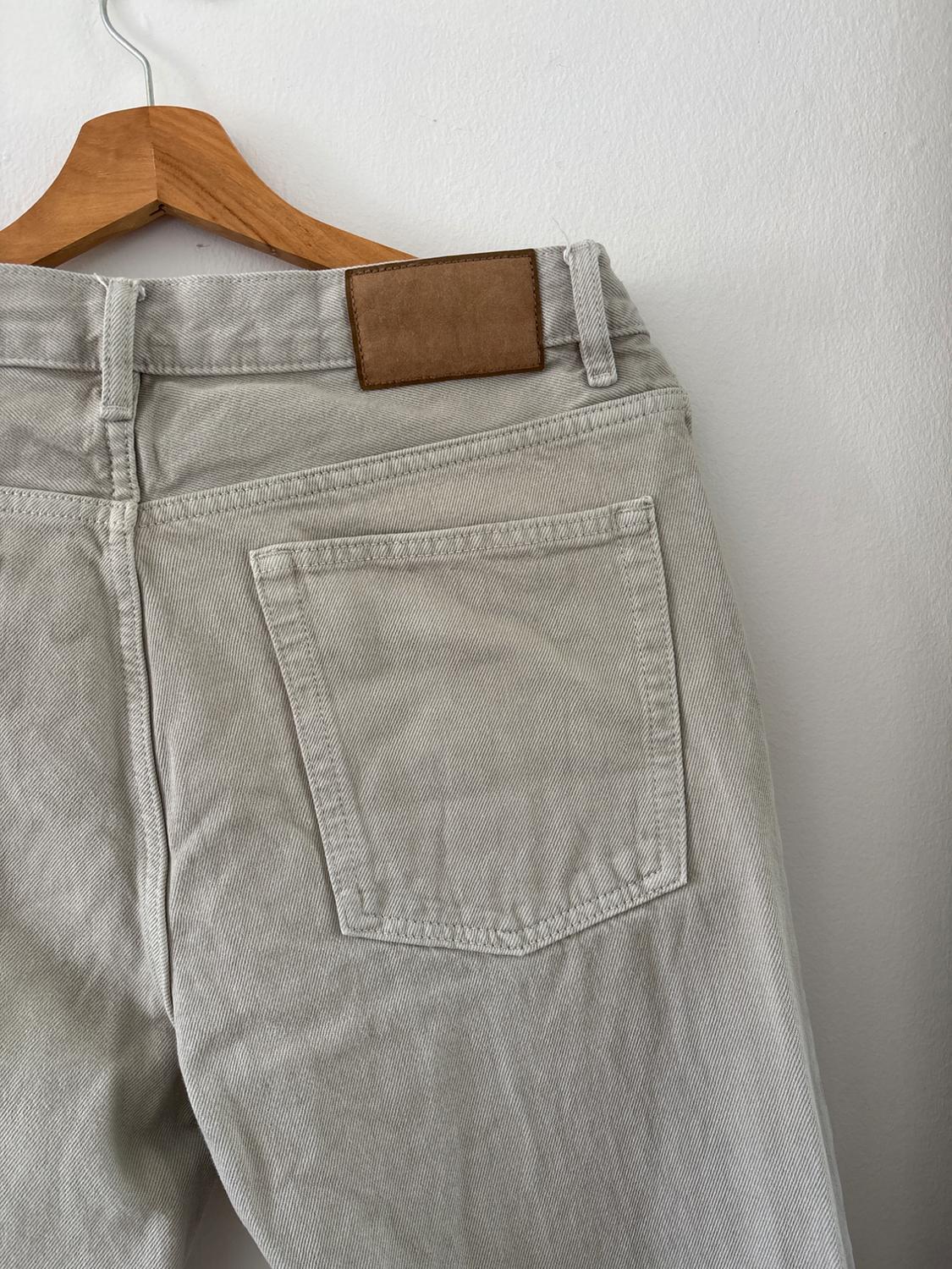 Back of light beige jeans hanging on a wooden hanger against a white wall. The pocket and a brown patch are visible.