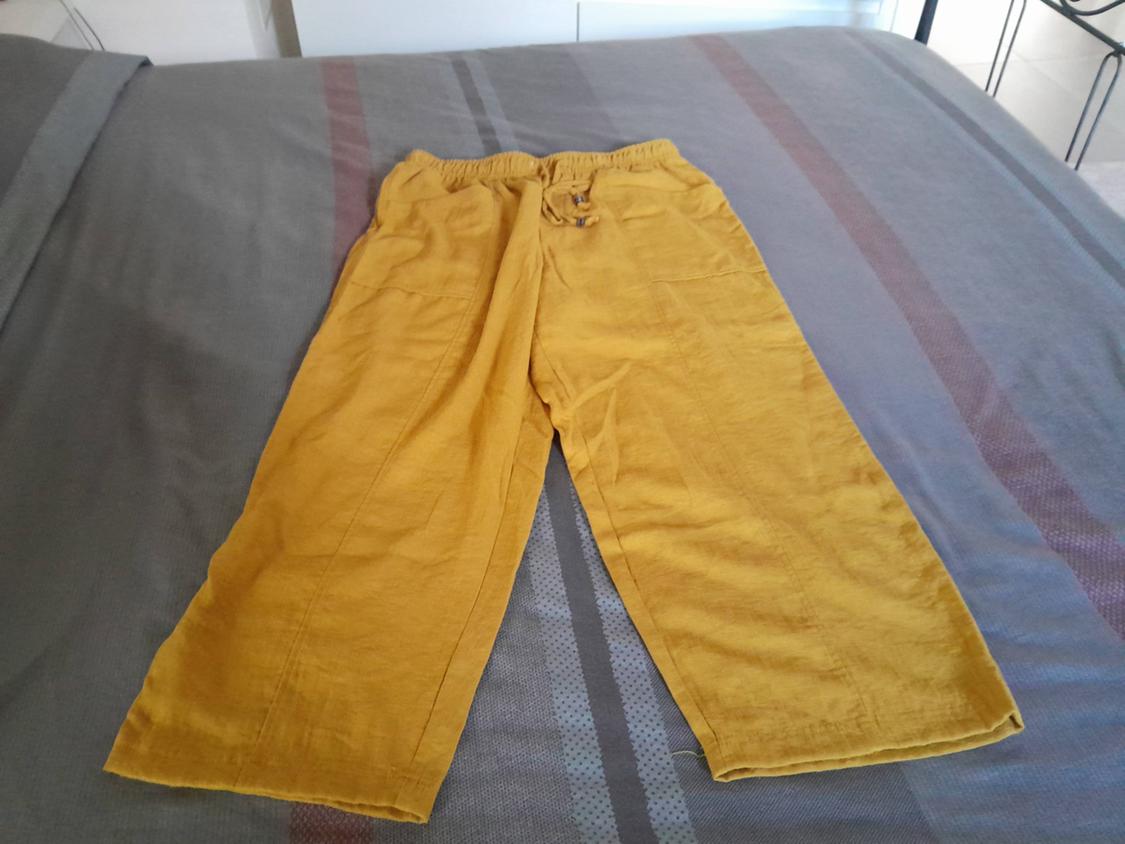 Yellow linen trousers with an elasticated waist, placed on a bed with a grey striped bedspread. Relaxed, homely atmosphere.