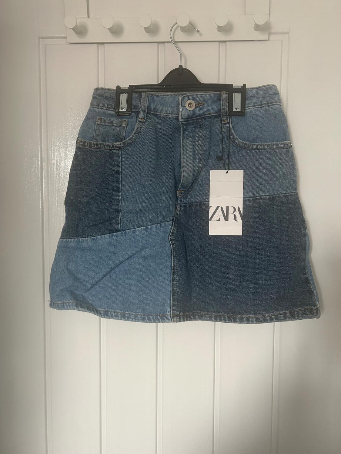 Short denim panelled skirt in various shades of blue, hanging on a hanger over a white door. Label with the text ZARA.
