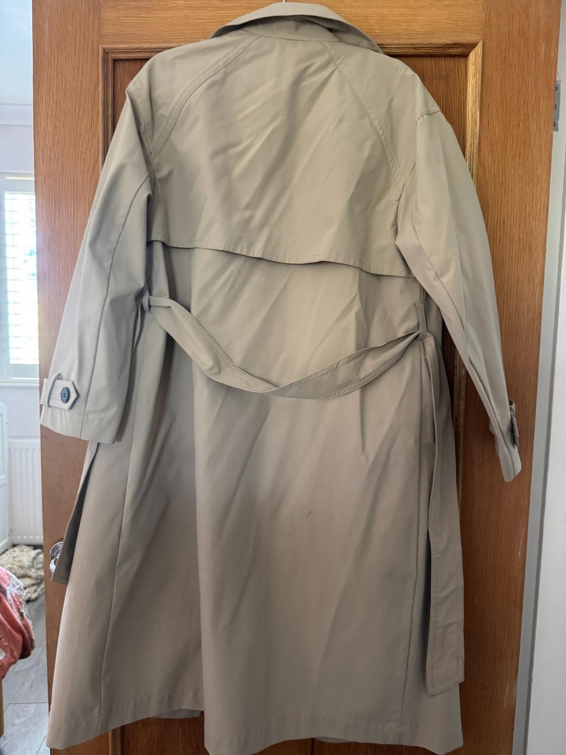 Beige trench coat seen from behind, hanging on a wooden door. Its knotted belt and back flap stand out in an indoor setting.