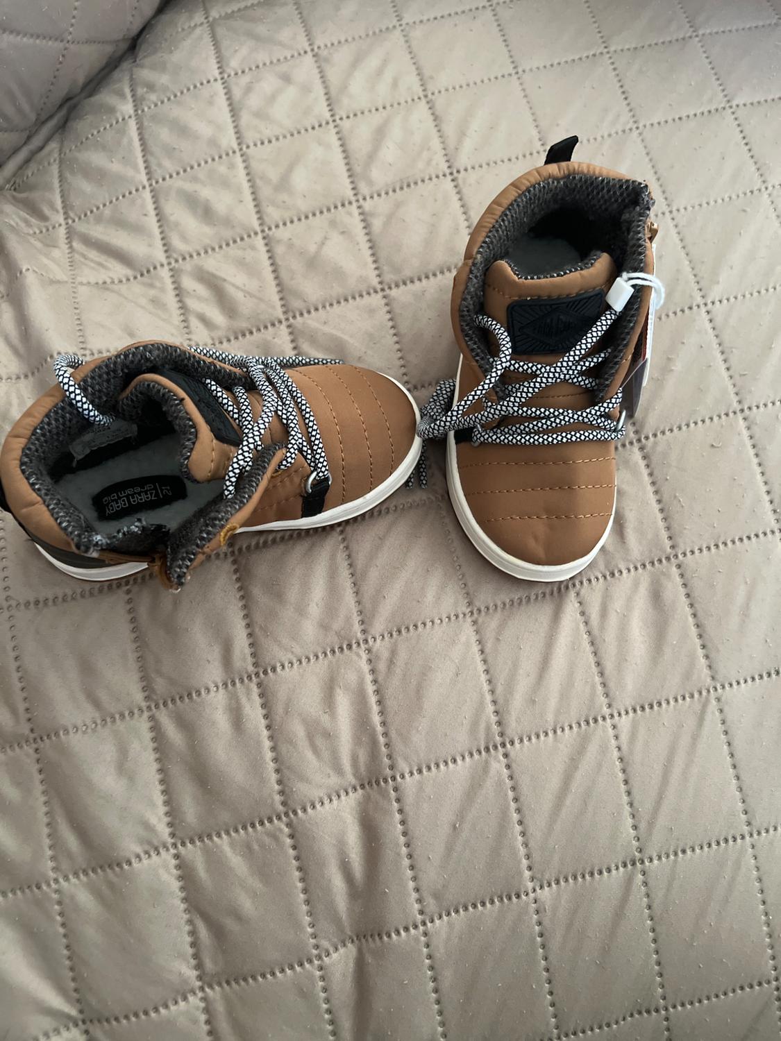 Brown boots with black and white laces on a beige quilted comforter. Visible text: "Zara Baby".