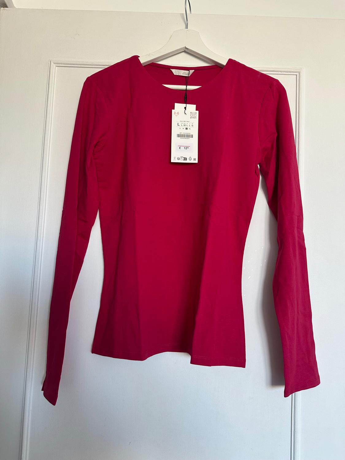 Red long-sleeved T-shirt hanging on a white hanger, with a visible label in the centre, against a white door background.