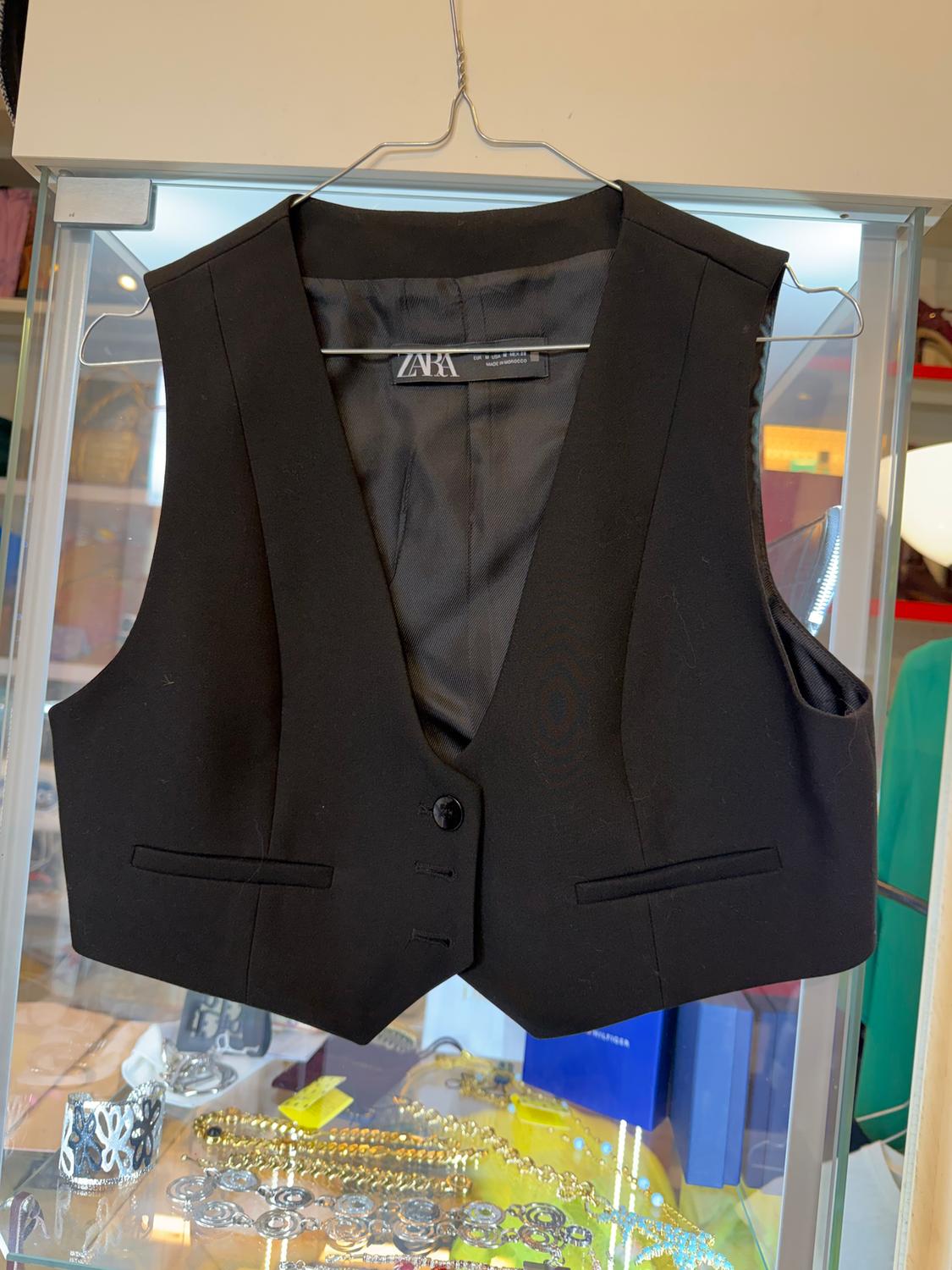 Black gilet from Zara hanging on a hanger inside a display case, with gold and silver accessories in the background.