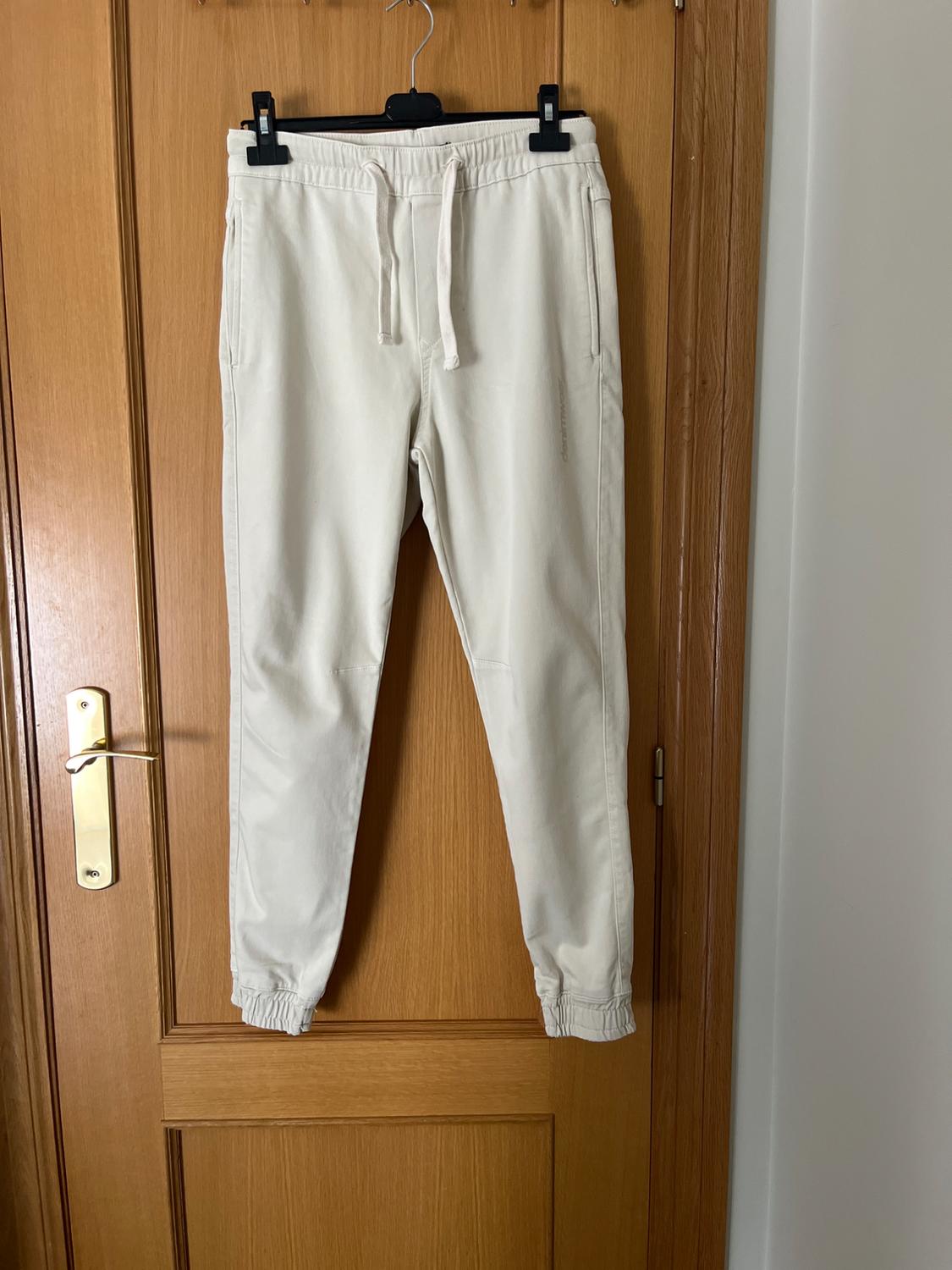 Beige jogger trousers hanging on a wooden door. Elastic drawstring waist and side pockets.