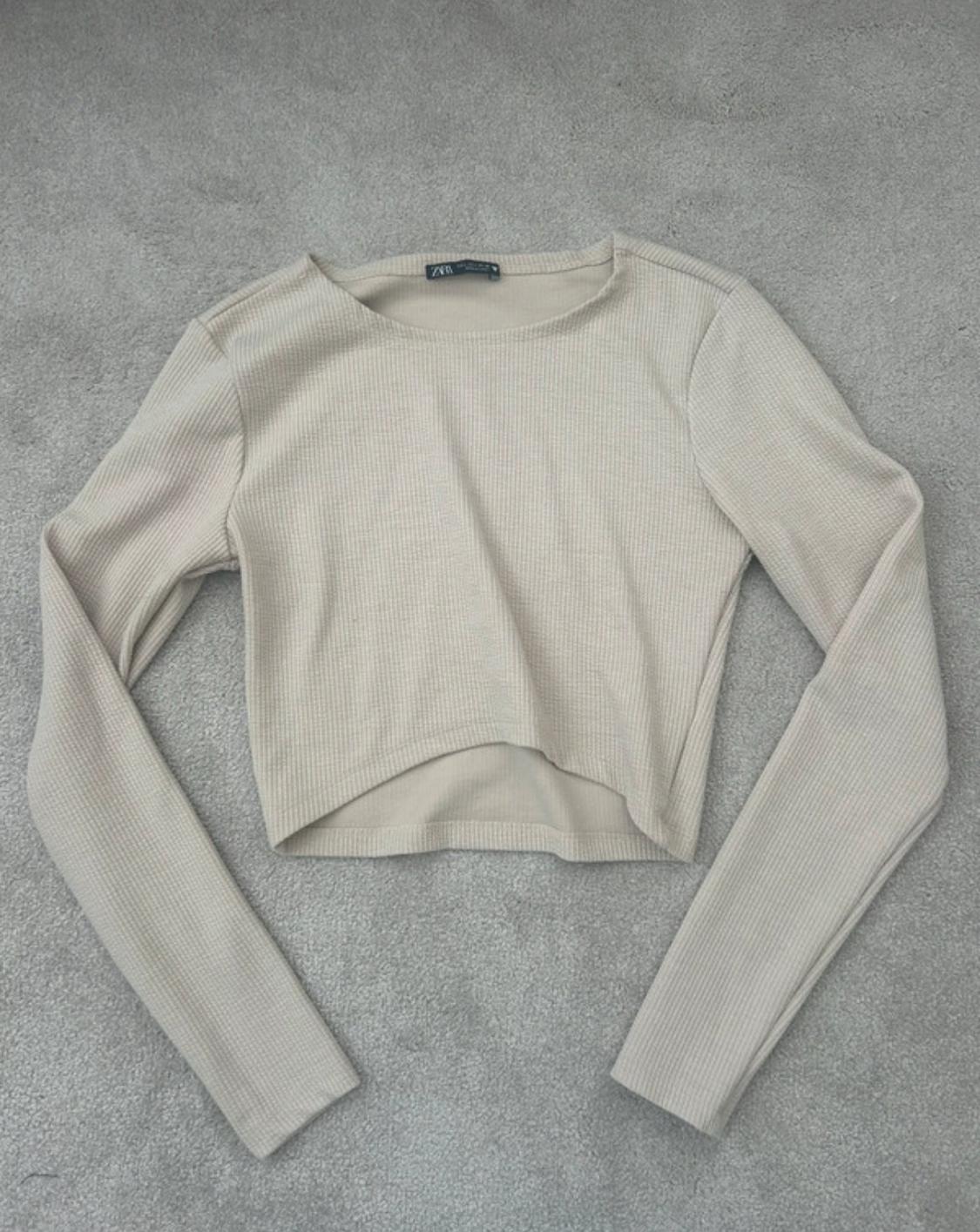 Beige long-sleeved crew-neck jersey on a grey rug. Visible label on the collar.