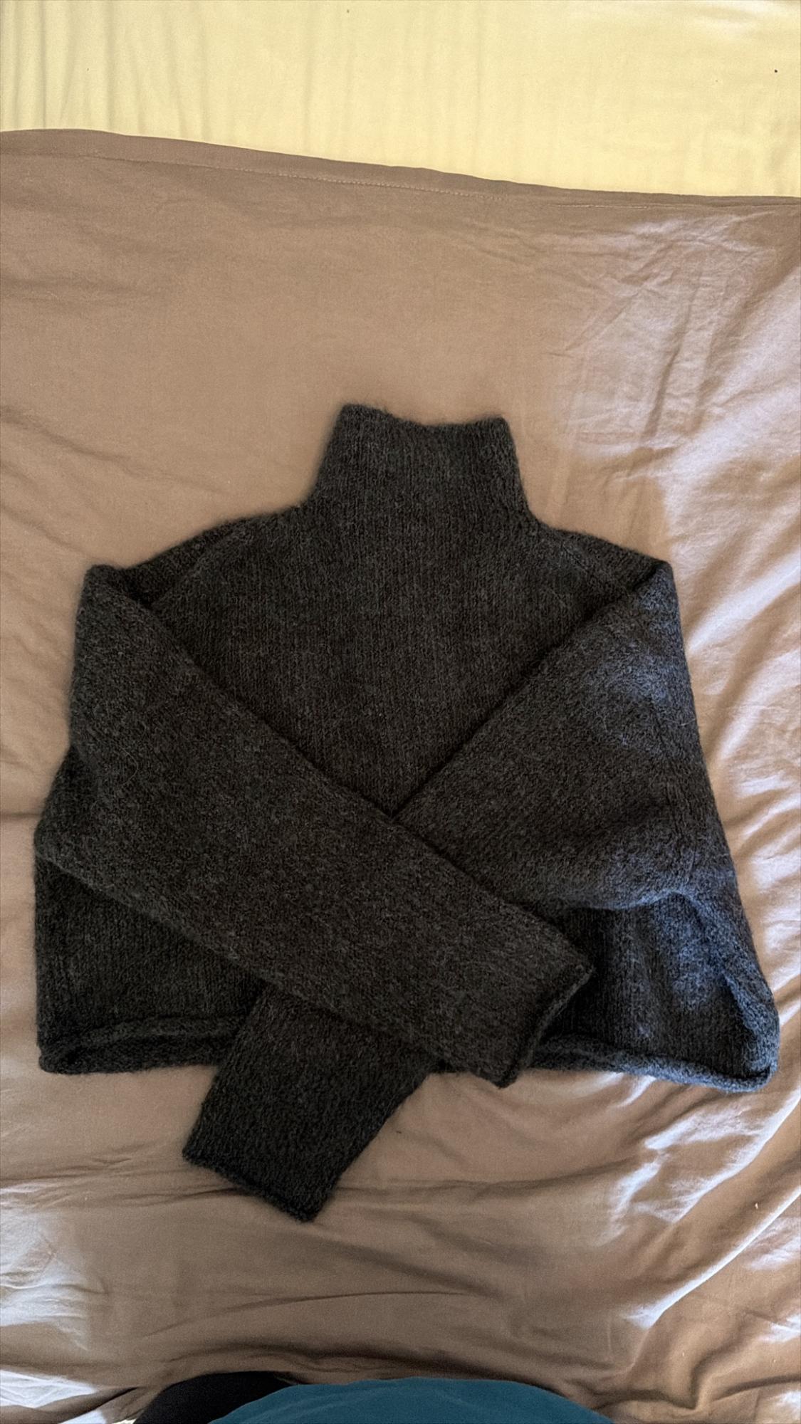 Dark grey knitted jumper with a high neck and cross-over sleeves, on a creased beige sheet. Casual and cosy style.