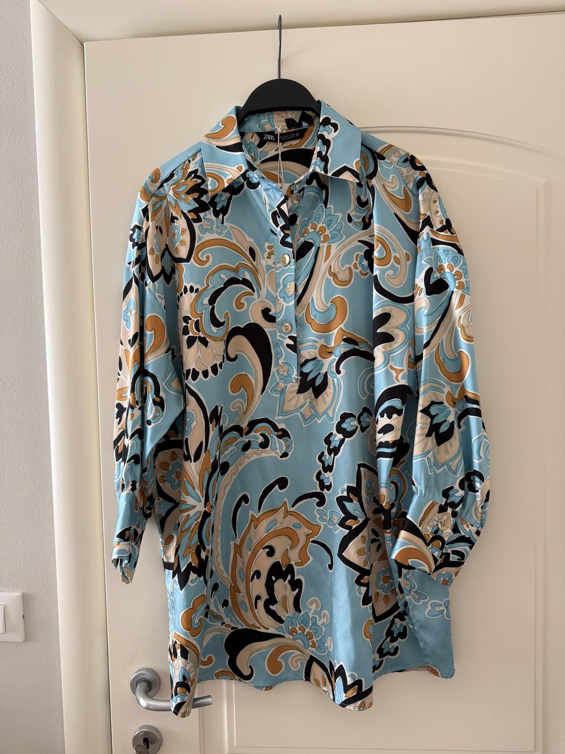 Silk shirt hanging on a white door, with a floral pattern in shades of blue, black, and beige. Visible brand label.