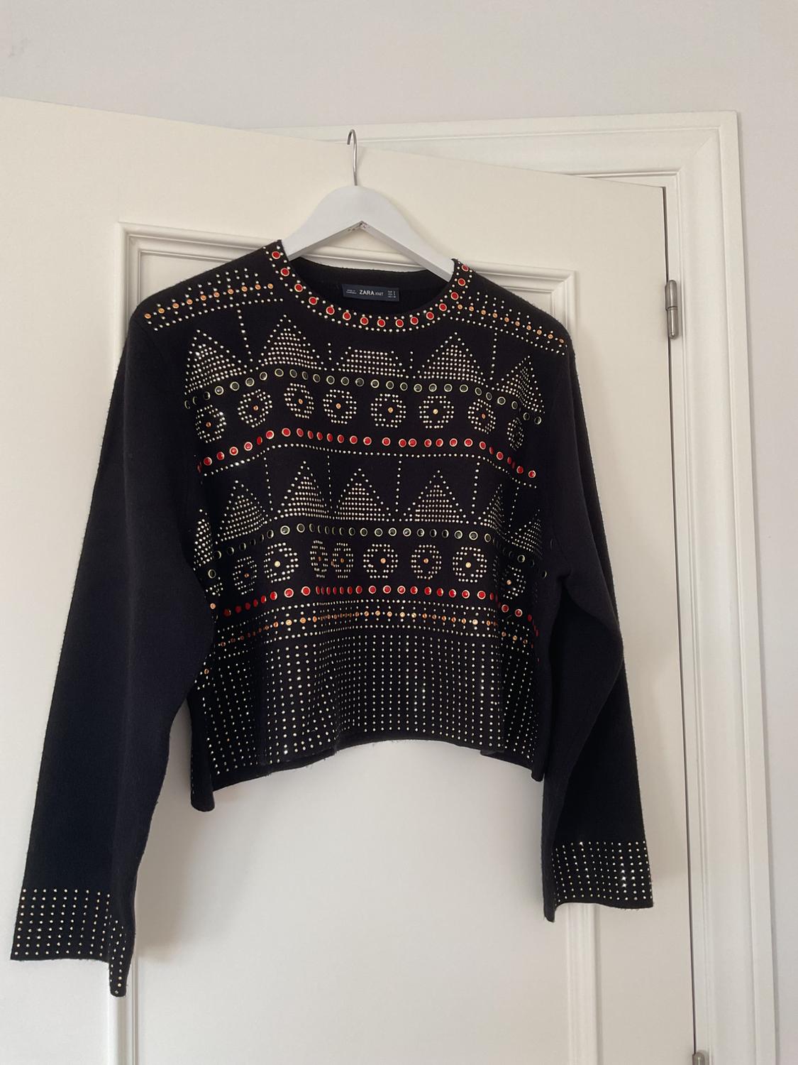 Black jumper with a geometric design of gold and red studs, hanging on a white hanger over a white door.
