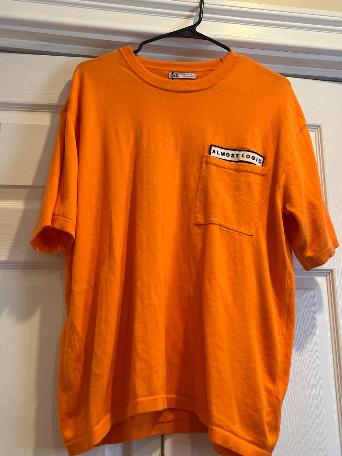 Orange t-shirt hanging on a hanger, with a chest pocket featuring the text "ALMOST LOGIC". White door in the background.