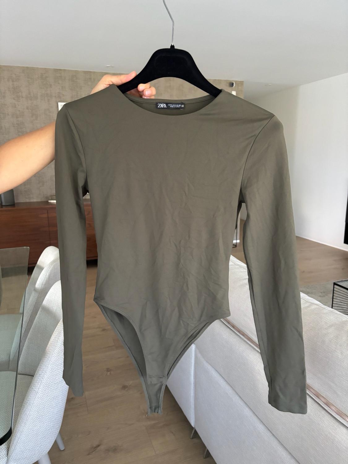 Arm holding an olive green long-sleeve bodysuit on a hanger. Living room background with a white sofa and table. Label with the text ZARA.