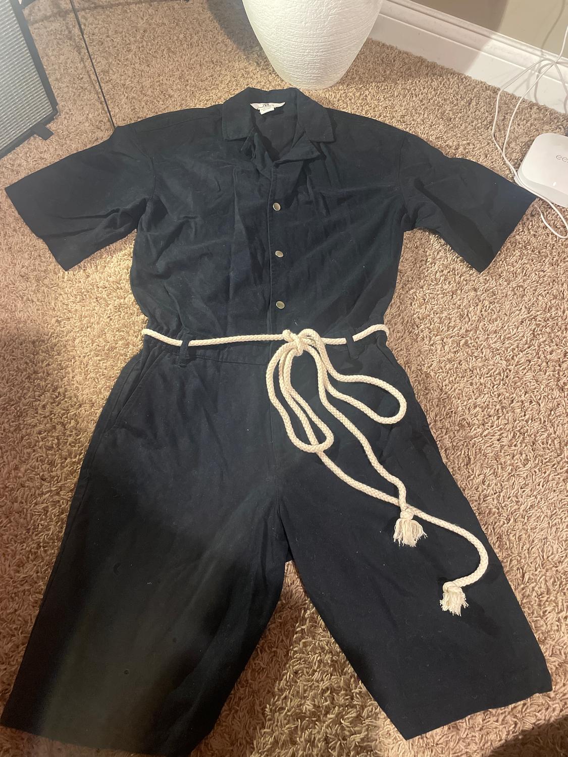 Black short-sleeved jumpsuit with buttons and a white rope belt, placed on a beige rug next to a white lamp.