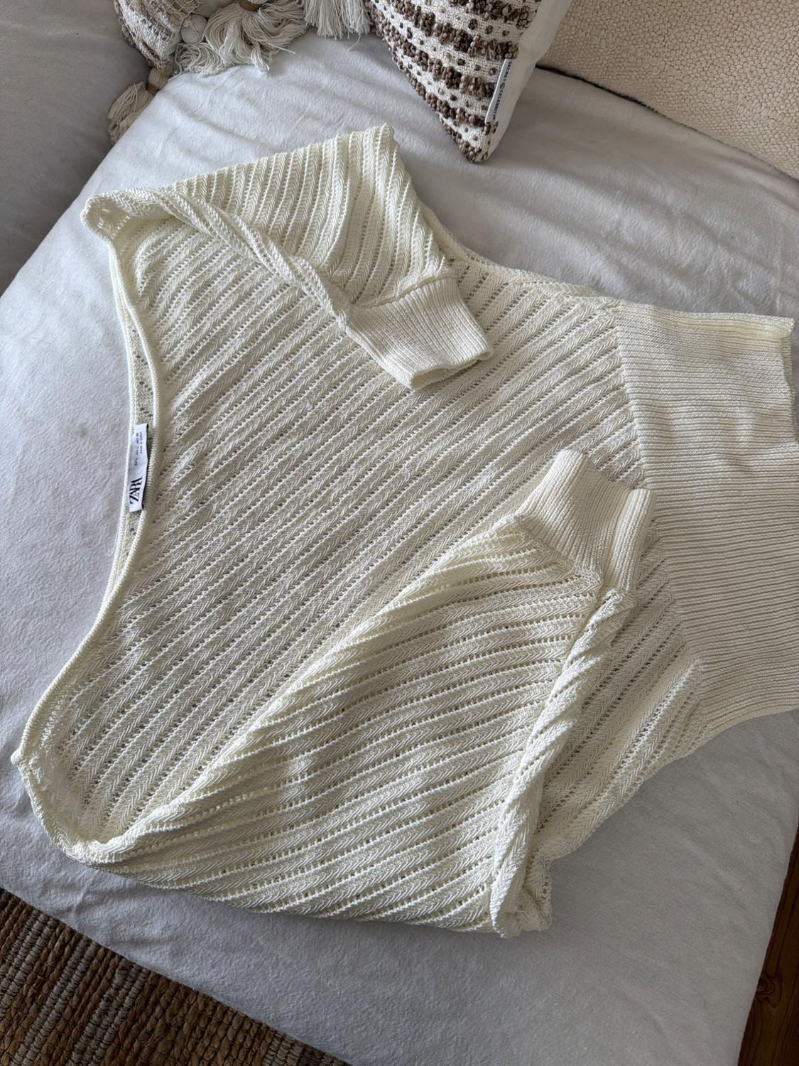 White knitted jumper with a diagonal texture and ZARA label, on a bed. Relaxed atmosphere with a decorative cushion in the background.