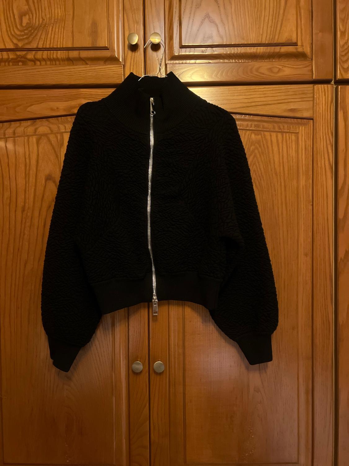 Black textured jacket with a silver zip and high neck, hanging in a wooden wardrobe.