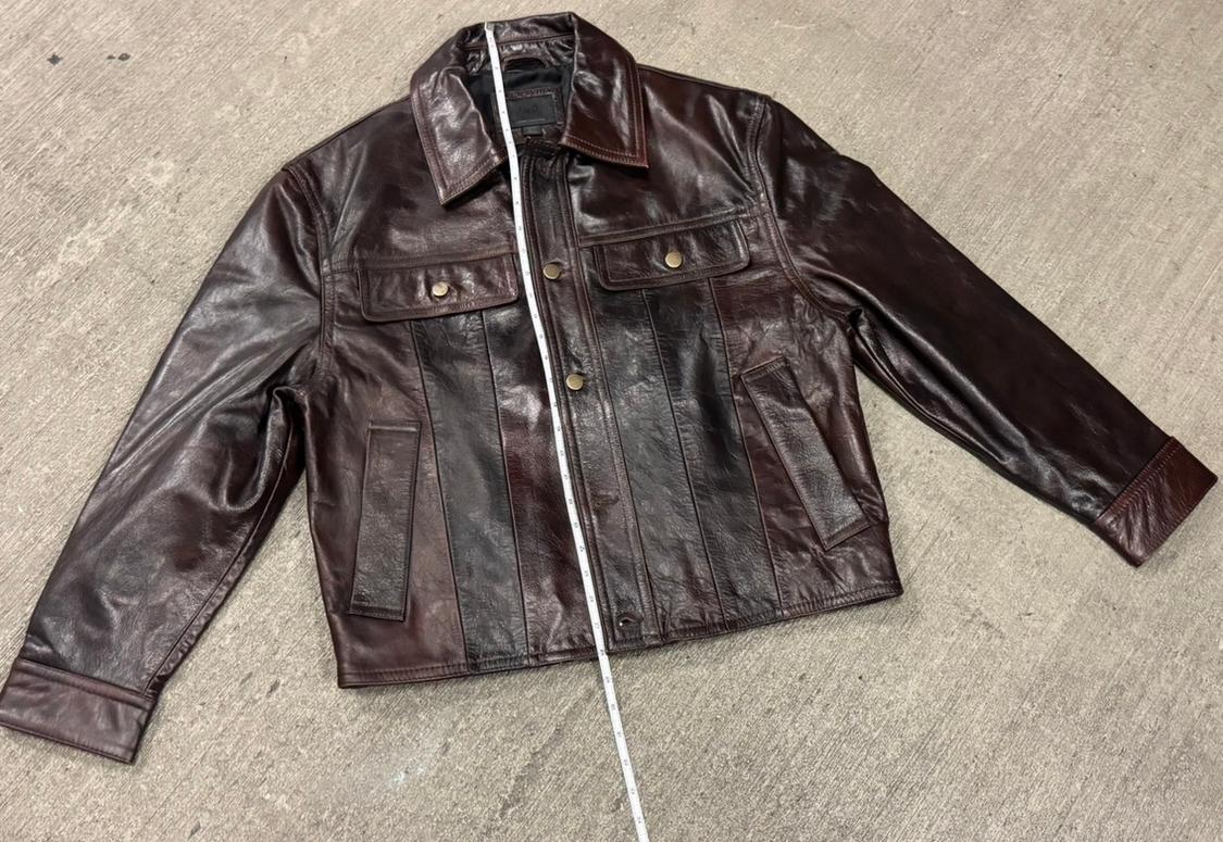 Dark brown leather jacket with front pockets and gold buttons, placed on a gray floor with a tape measure on top.