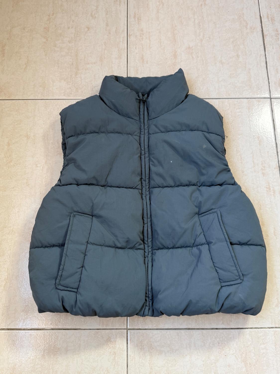 Dark grey padded gilet on a light tiled floor. Sleeveless design with front pockets and front fastening.