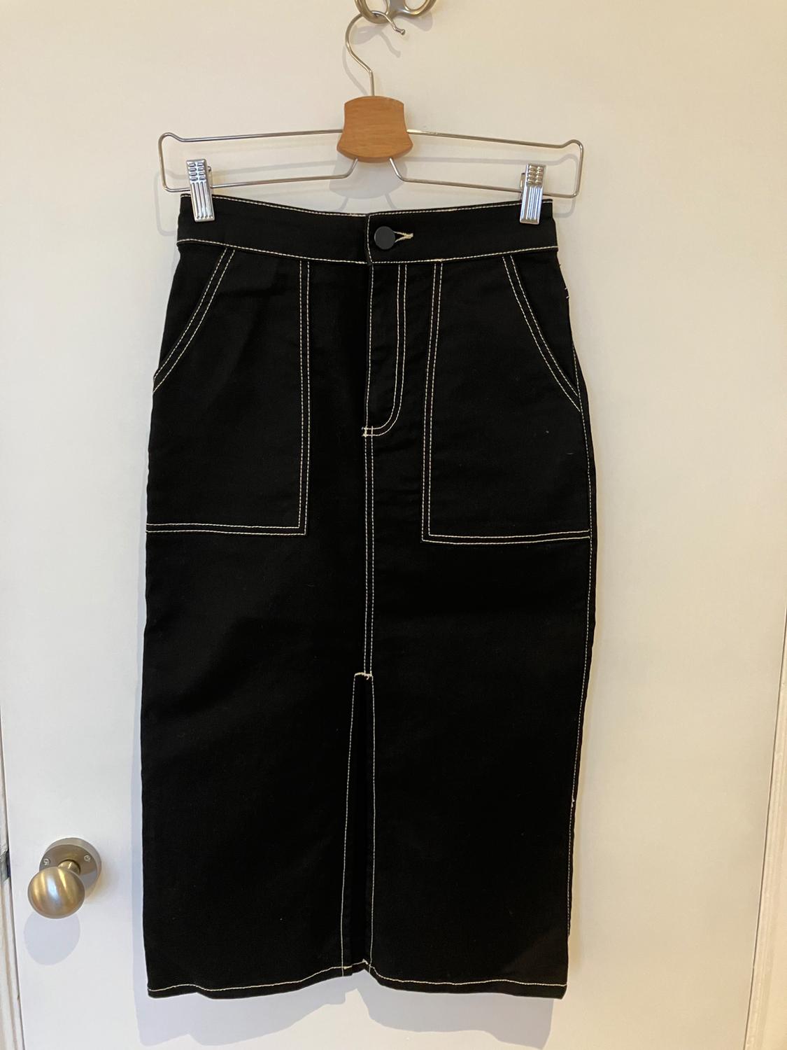 Black denim skirt with visible white stitching, large front pockets, hanging on a wooden hanger over a white door.