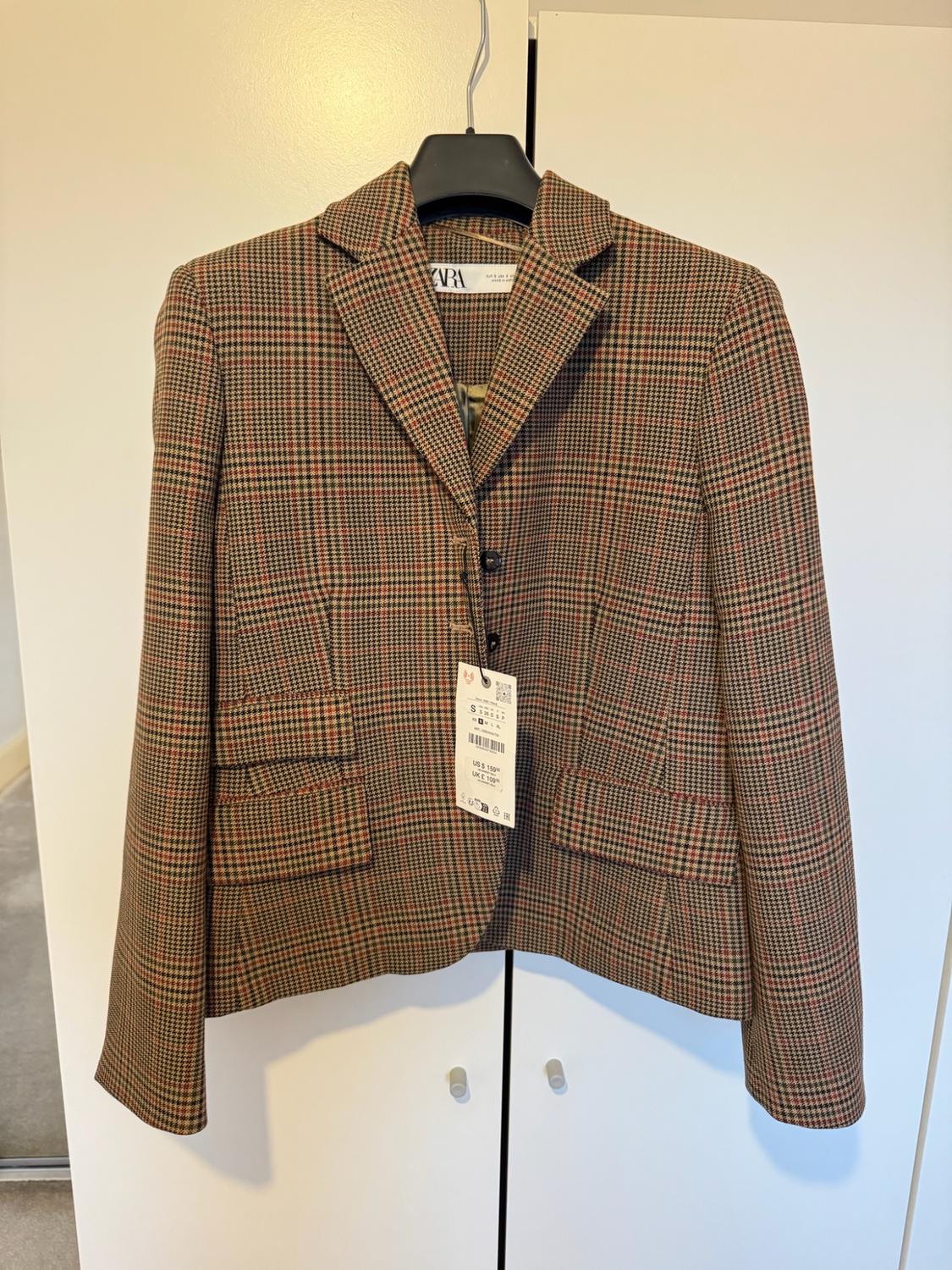 Brown checked blazer hanging on a hanger in front of a white wardrobe. Label on the collar with the text ZARA.