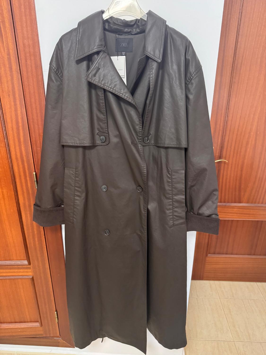 Dark brown long coat hanging on a hanger between two wooden doors. Its buttons and a label with the text ZARA stand out.