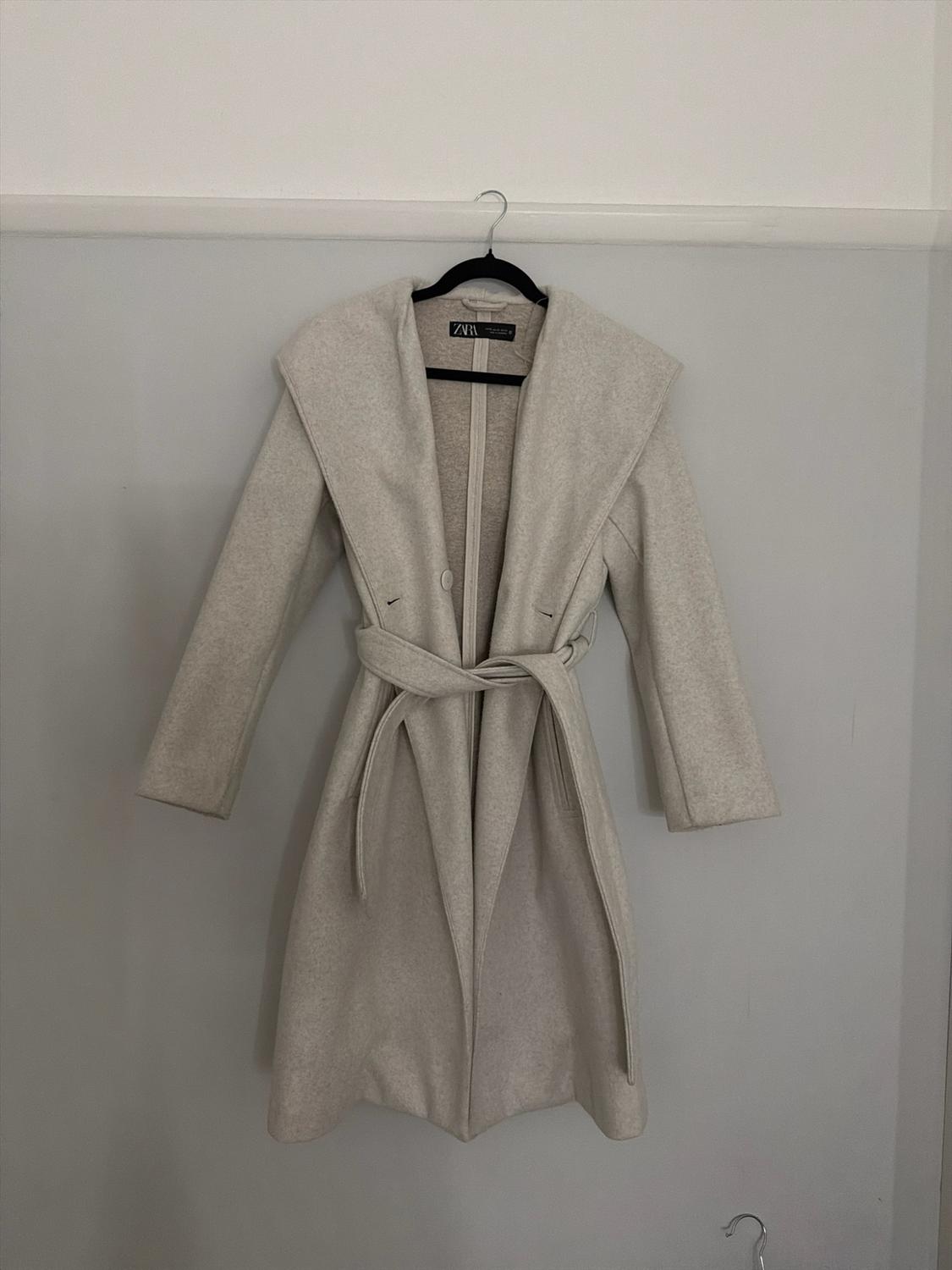 Light beige coat with a wide collar and tied belt, hanging on a black hanger against a light wall. ZARA label visible.