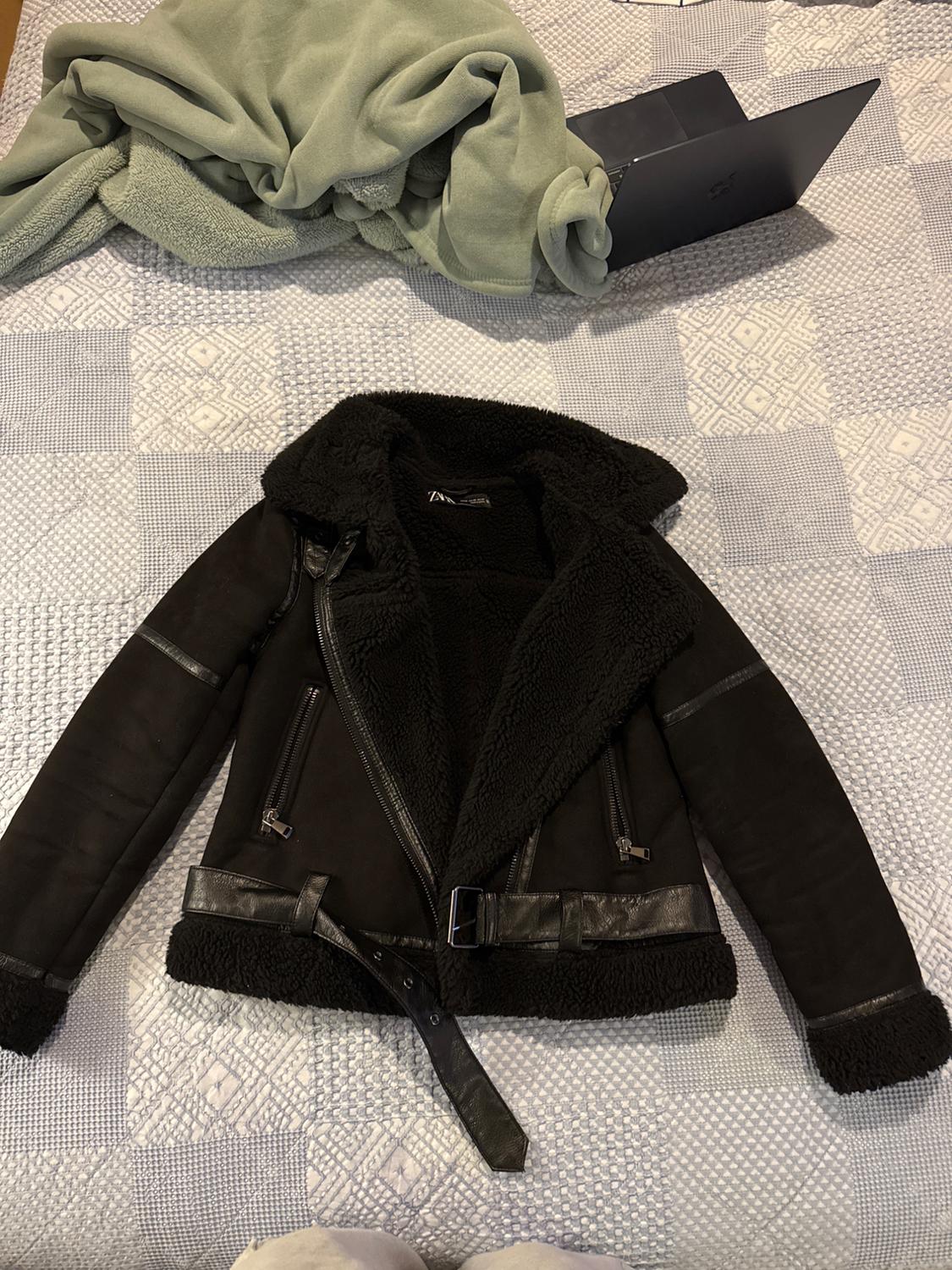Black jacket with a sheepskin lining and a belt on a bed with a gray plaid comforter. Green blanket and laptop in the background.
