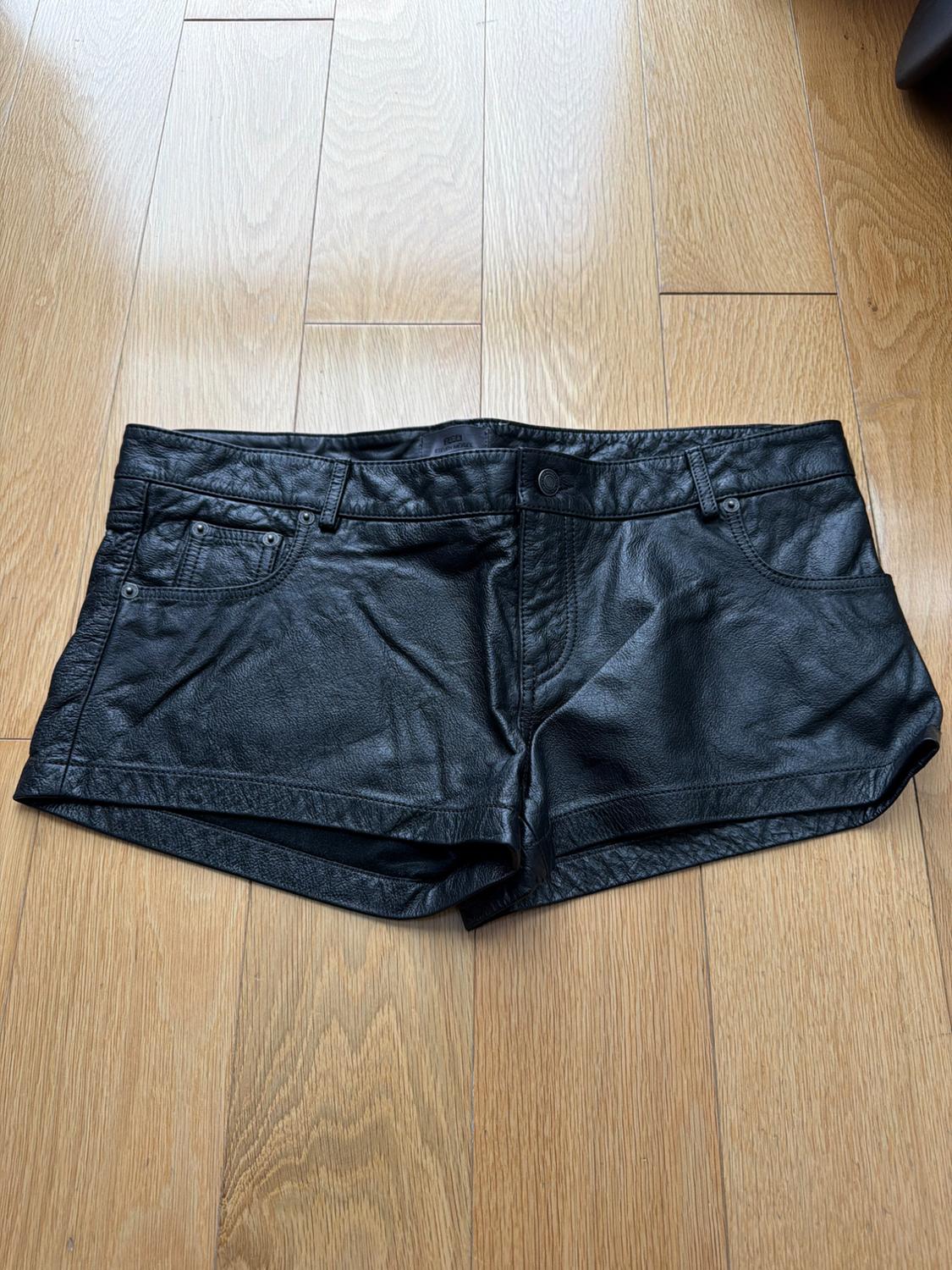 Black faux leather shorts on a light wood floor. Design with front pockets and belt loops.