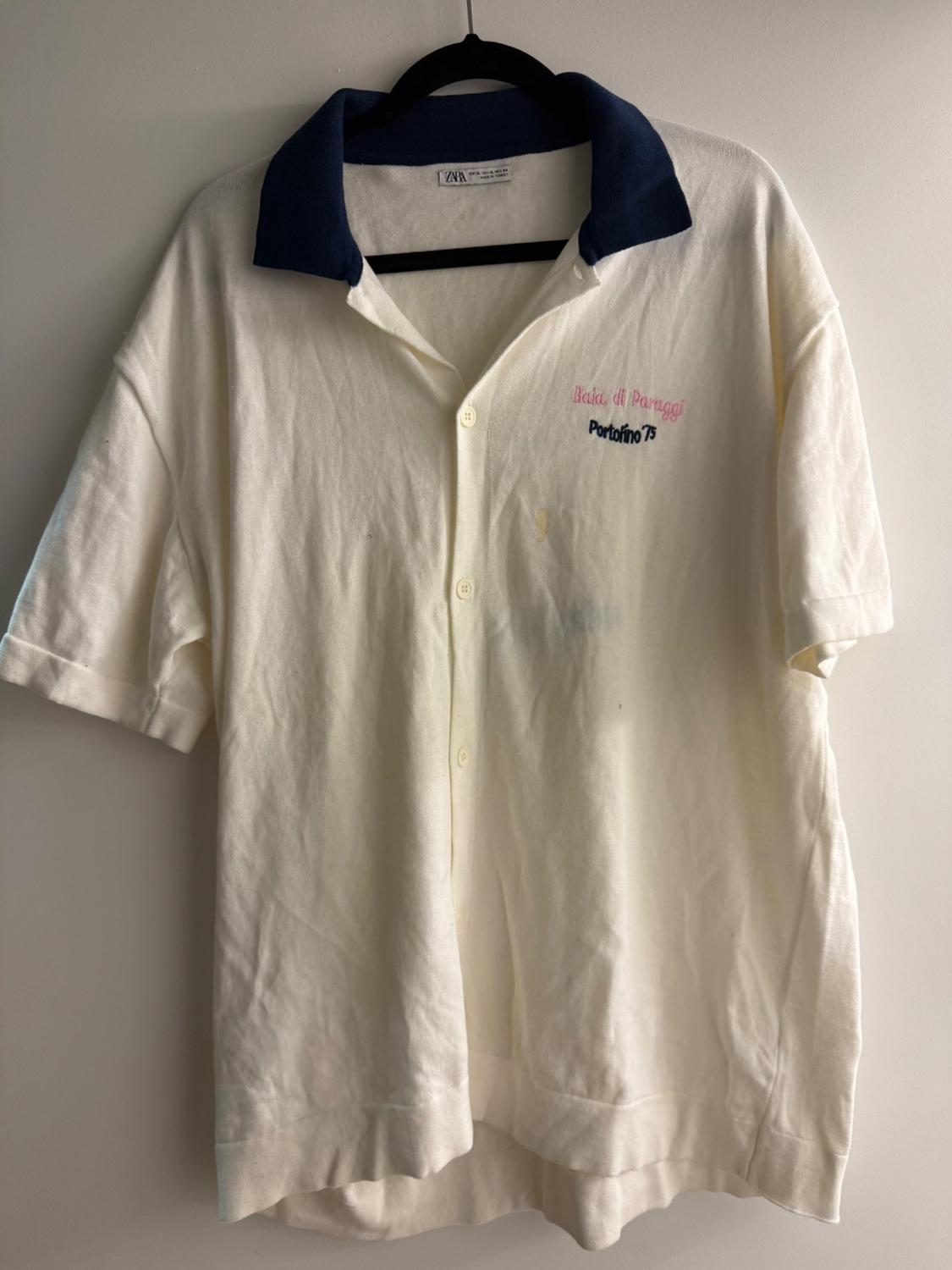White short-sleeved shirt with navy blue collar. Embroidered text: "Baia di Paraggi Portofino 75". Hung on a black hanger.