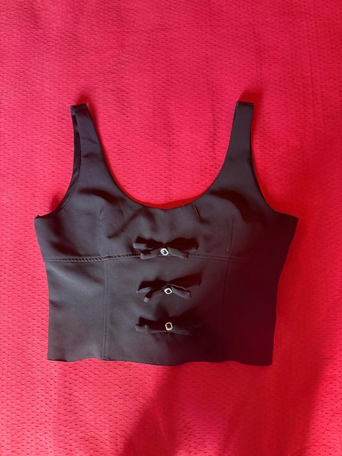 Black sleeveless top with three front bows and shiny details, displayed against a textured red fabric background.