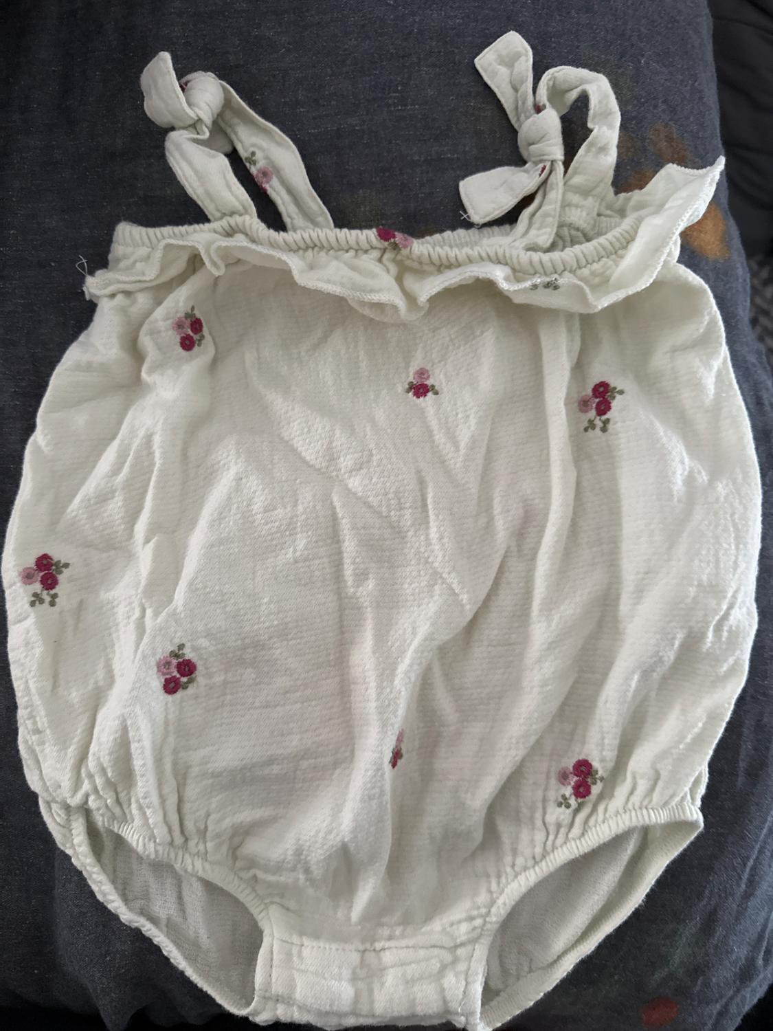 White romper with tied straps and pink and red floral embroidery on a dark background.