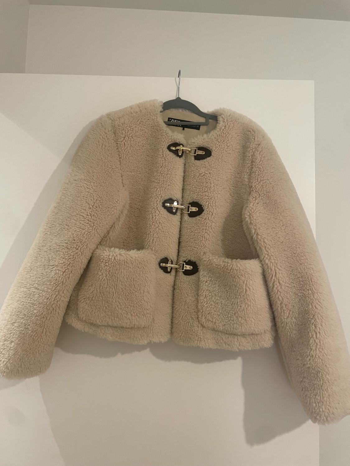 Beige faux fur jacket on a hanger. It features three metal fastenings with dark details, front pockets and a ZARA label.