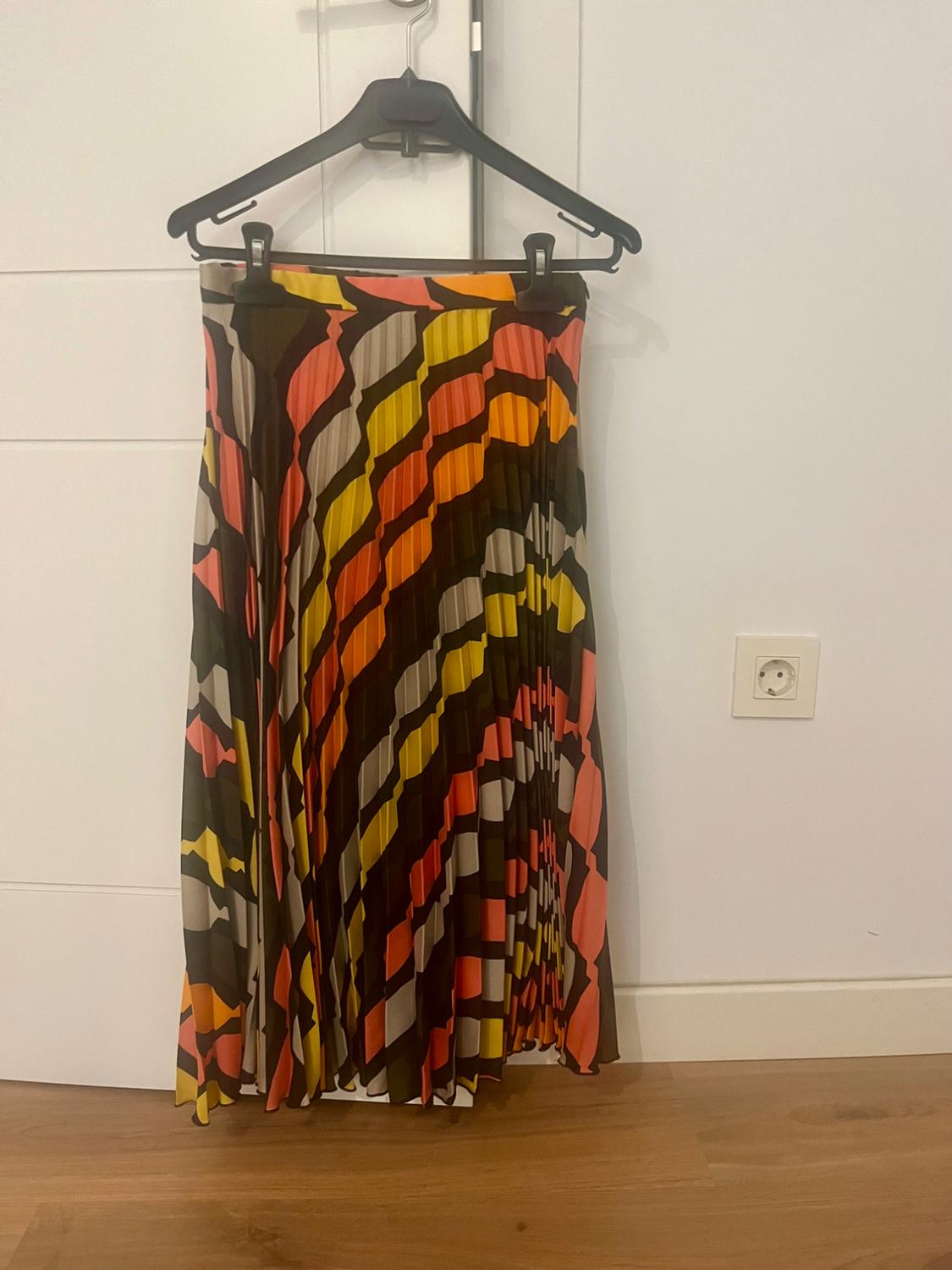 Pleated skirt with geometric print in shades of orange, yellow and black, hanging on a black hanger over a white door.