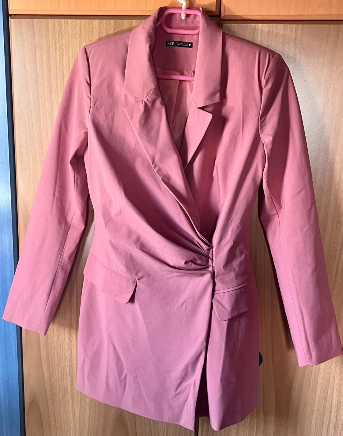 Pink blazer with a lapel collar and draped detailing, hanging on a hanger in front of a wooden wardrobe. Label with the text ZARA.